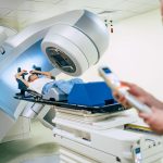 A patient lies beneath a radiation treatment machine while a clinician operates its controls