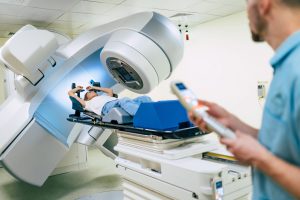 A patient lies beneath a radiation treatment machine while a clinician operates its controls