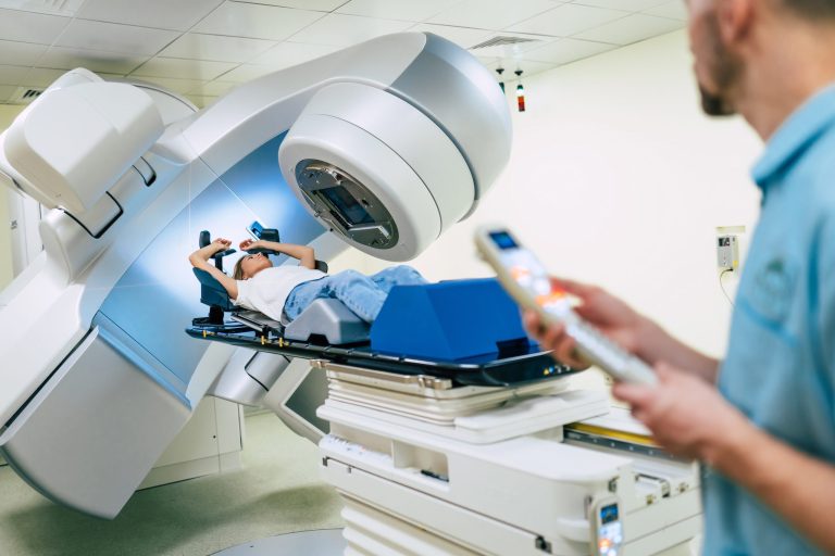 A patient lies beneath a radiation treatment machine while a clinician operates its controls