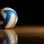 A blue and white volleyball rests on a wooden floor against a dark background