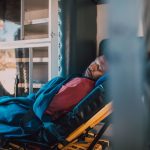 An adult man rests on a stretcher under a blue blanket