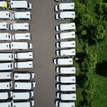 White buses are parked in rows beside a tree-lined lot